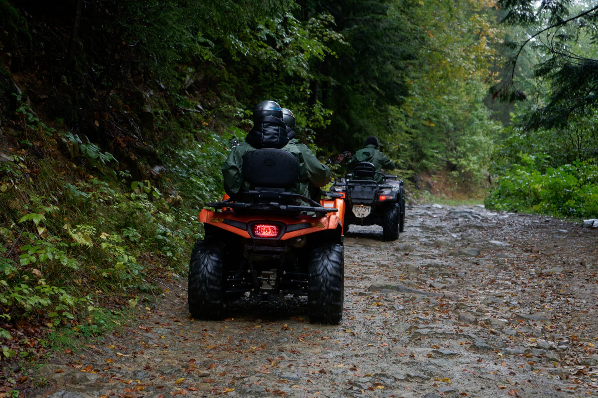 Two ATV riders on a wet gravel forest trail